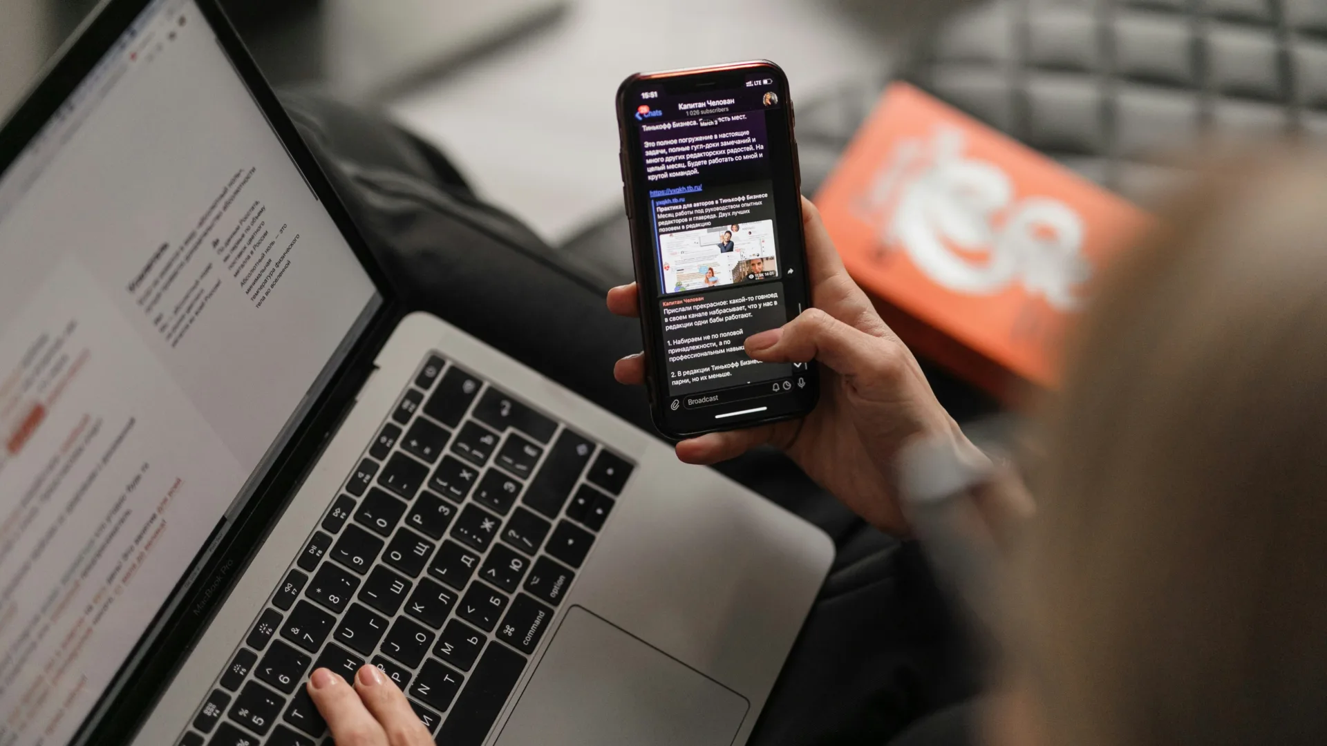 View over a person's shoulder as they work on a laptop while simultaneously viewing news on their phone.